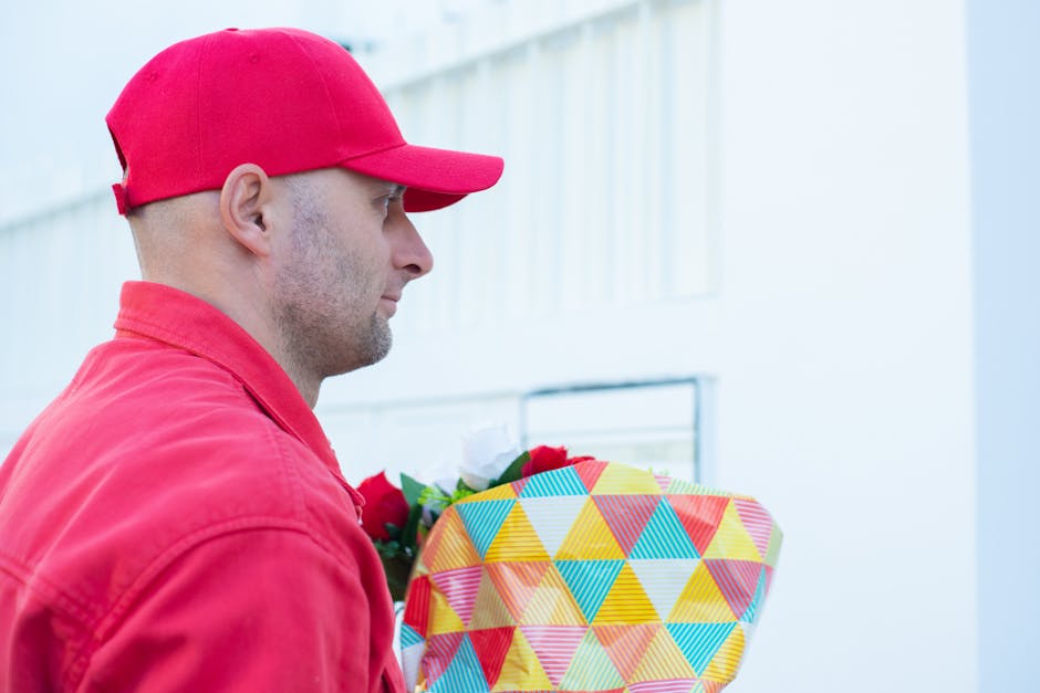 A delivery person wearing a red cap and matching red jacket holds a colorful floral gift wrapped in a geometric patterned paper with shades of yellow, blue, red, and green. The person faces to the lef